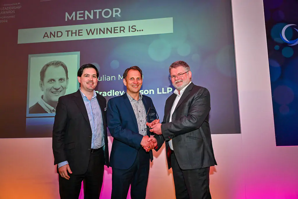 Three men stand together on stage, with one receiving an award during a ceremony.