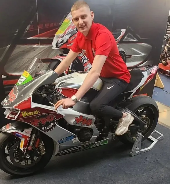 A young man in a red shirt sits on a racing motorcycle indoors.